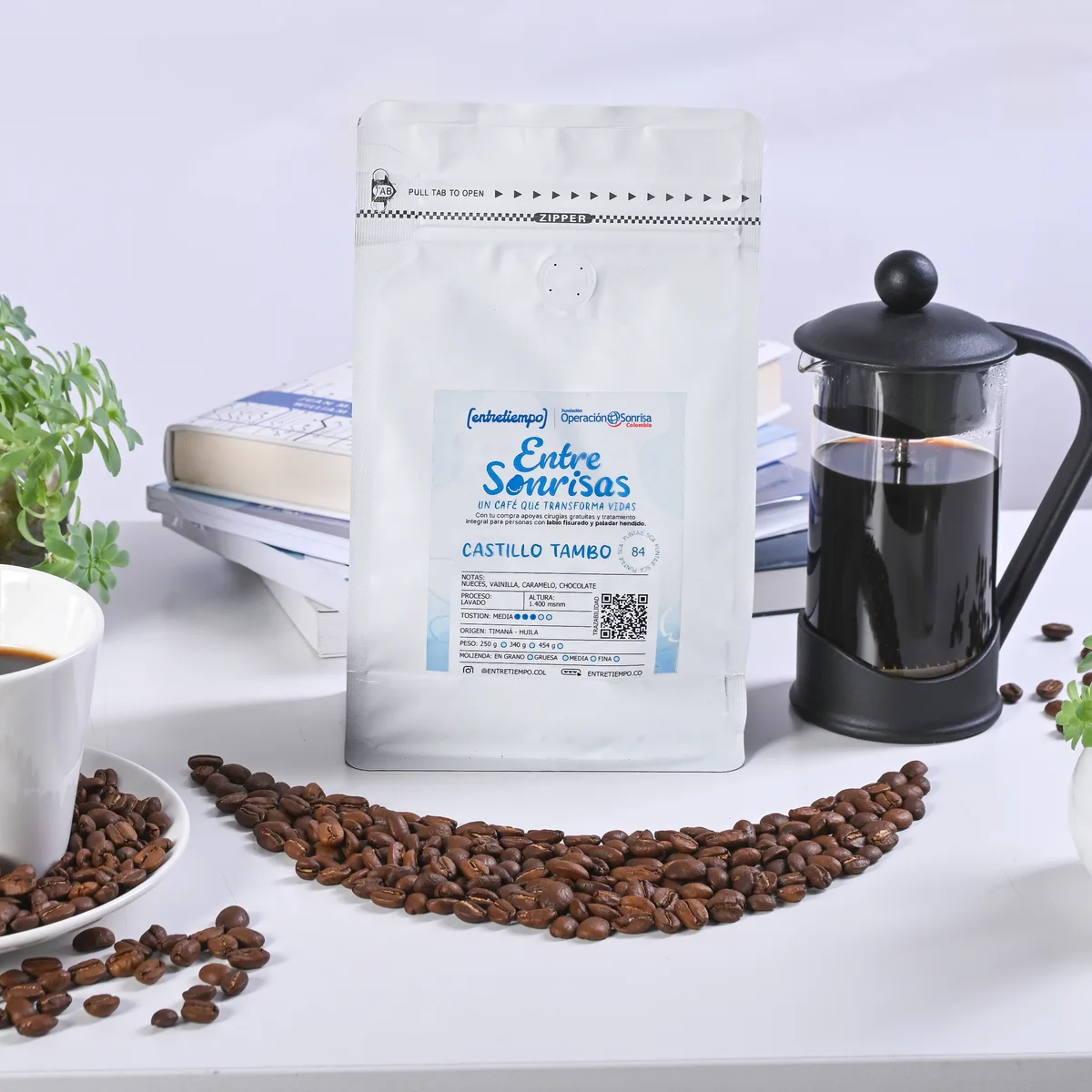 Bolsa del café Entre Sonrisas sobre una mesa junto a una taza de café, una prensa francesa y granos de café.